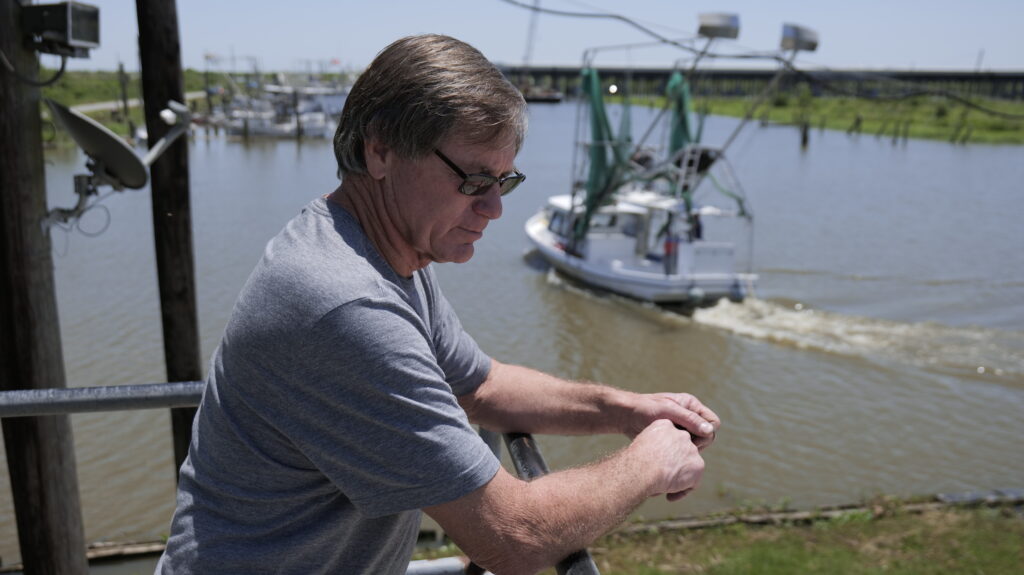 Don Beshel on the balcony of his marina near Point á la Hache, as a shrimp boat leaves one morning in April 2022. Beshel’s Marina is near Mardi Gras Pass, a breach in the Mississippi River levee that occurred naturally in 2011.