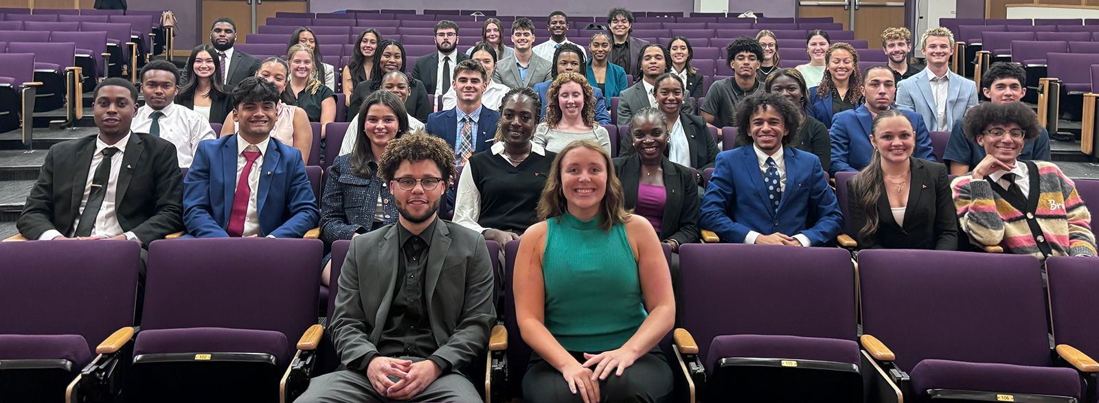 Students wear professional attire and sit in purple seats in the BEC auditorium. They smile warmly as they look directly at the camera. 