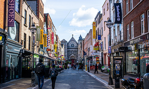 Pedistrians walk up a shopping street in Dublin, a church appears in the background.
